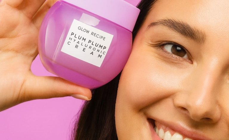 Joven con piel radiante sosteniendo Glow Recipe Plum Plump Hyaluronic Cream frente a fondo rosa pastel.