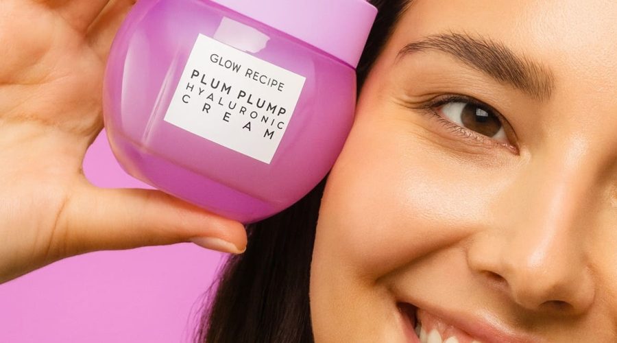 Joven con piel radiante sosteniendo Glow Recipe Plum Plump Hyaluronic Cream frente a fondo rosa pastel.